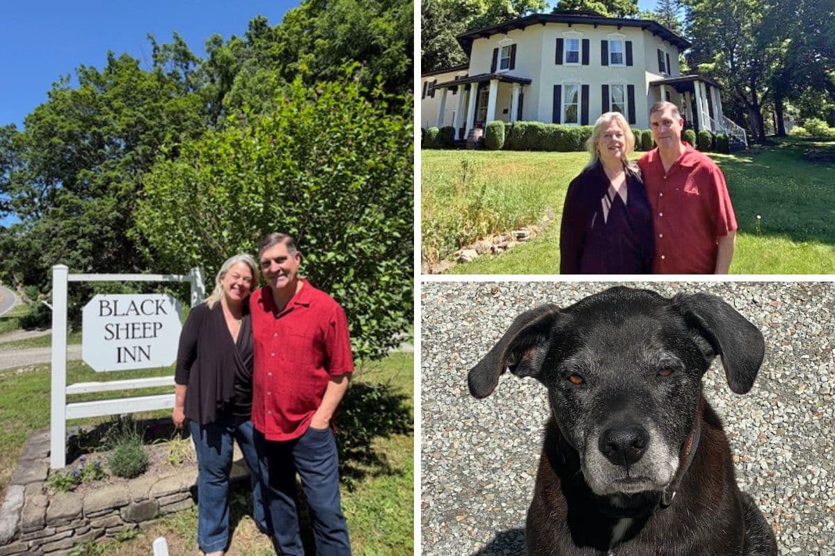 A couple stands by a "Black Sheep Inn" sign, with a dog and a view of the inn in the background.