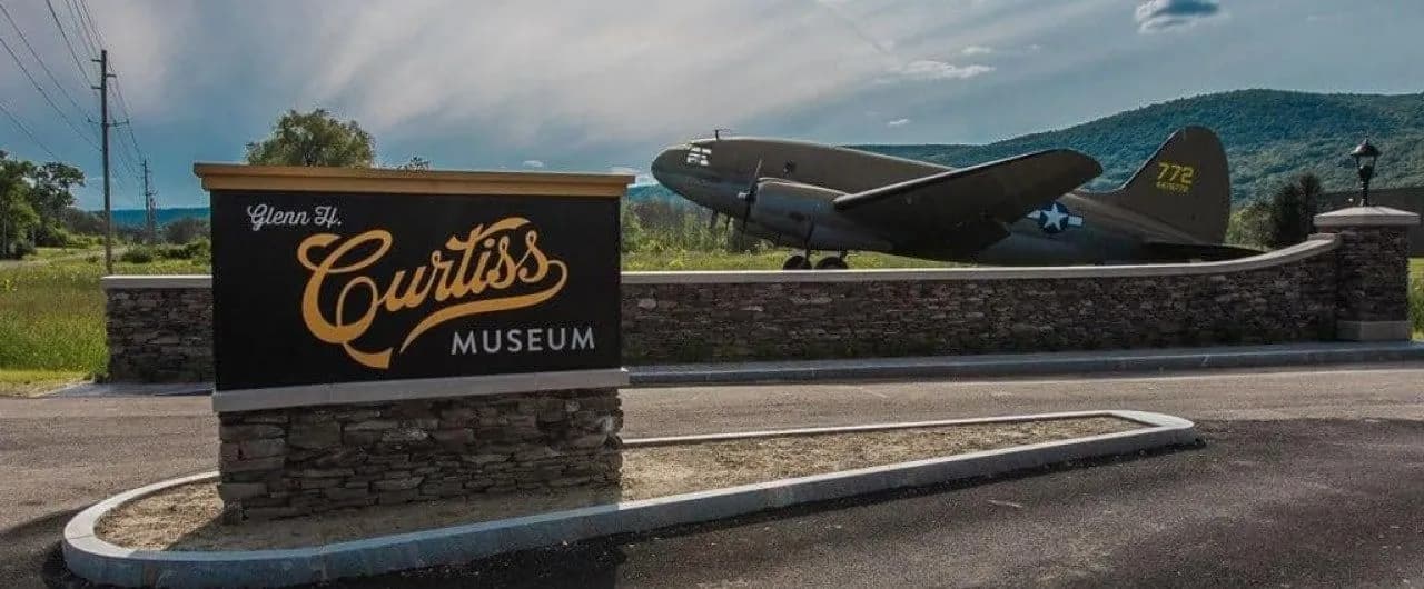 Sign for the Glenn H. Curtiss Museum with a vintage airplane in the background.