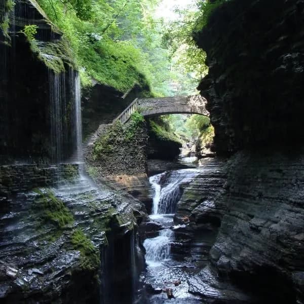 A serene gorge featuring cascading waterfalls and a stone bridge surrounded by lush greenery.