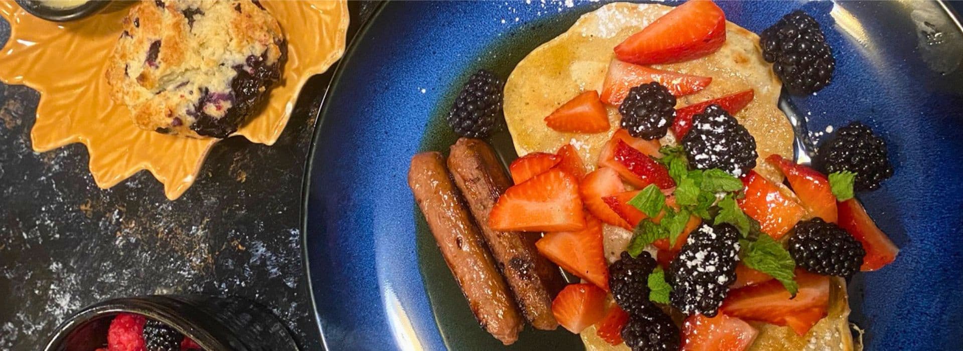 A plate of pancakes topped with strawberries, blackberries, and mint, accompanied by sausage links, with a muffin and berries on the side.