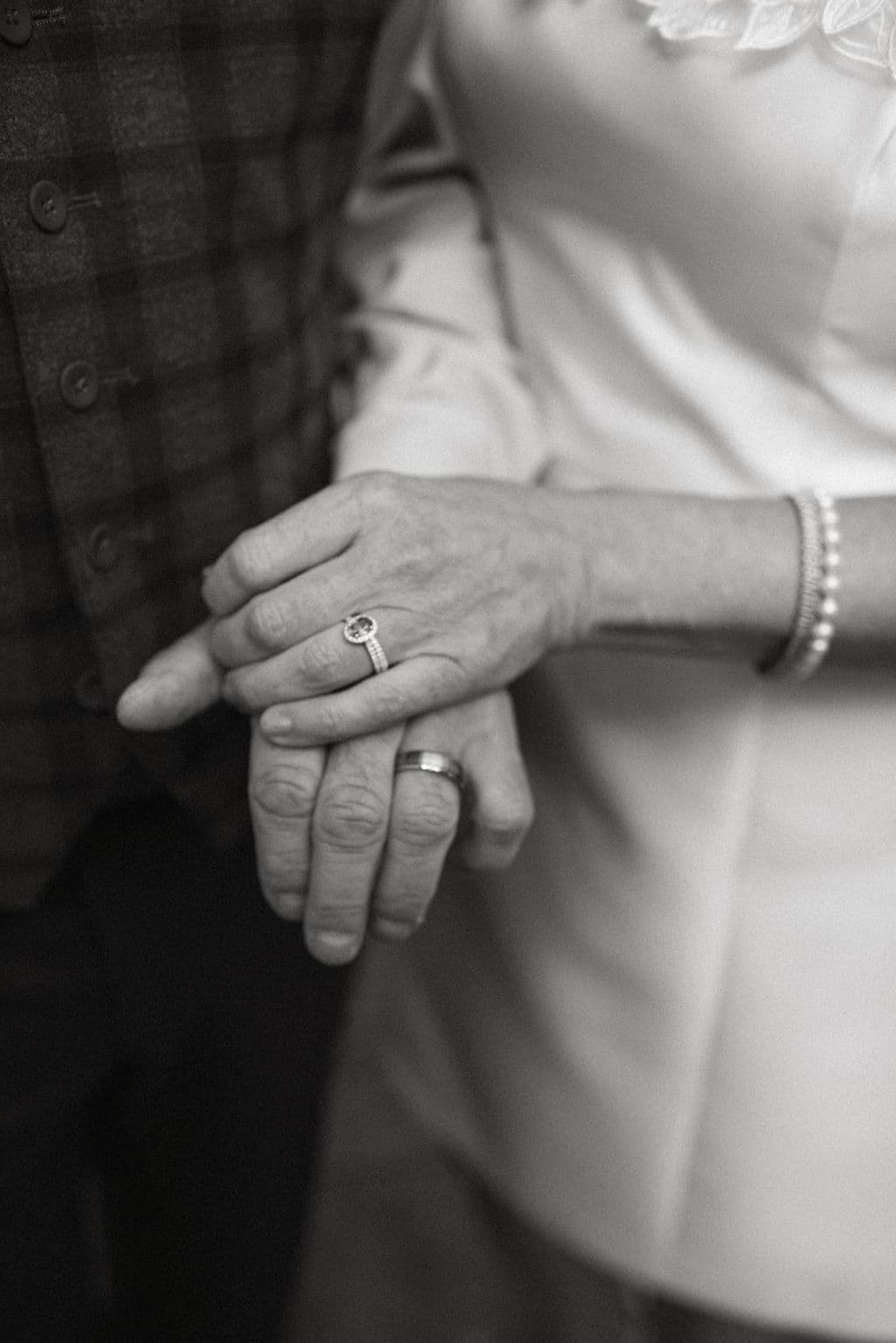 A close-up of two hands intertwined, one with a diamond ring and the other wearing a simple band.