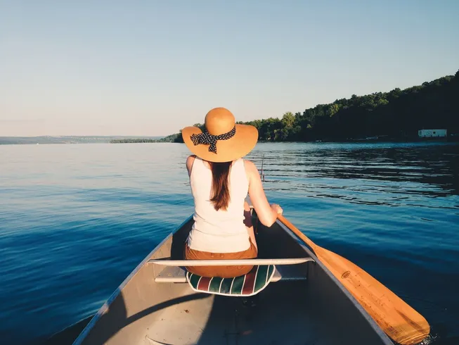 A woman in a wide-brimmed hat rows a canoe on a calm lake.