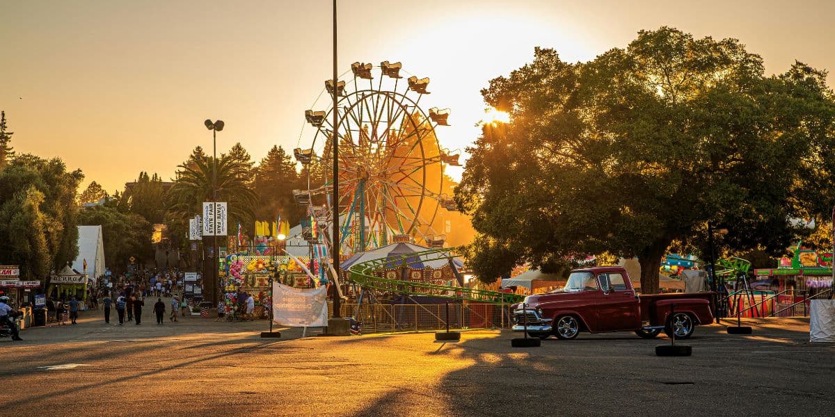 Sunset over a carnival with a Ferris wheel and classic truck parked nearby.