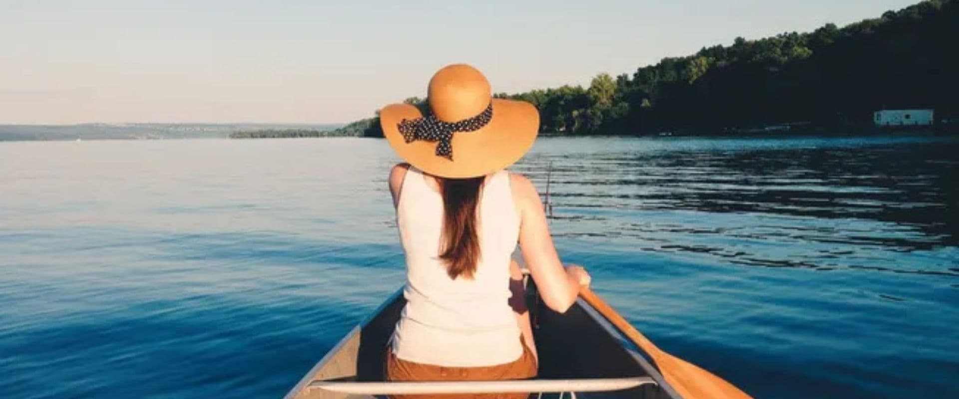 A person in a wide-brimmed hat paddles a canoe on a calm lake.