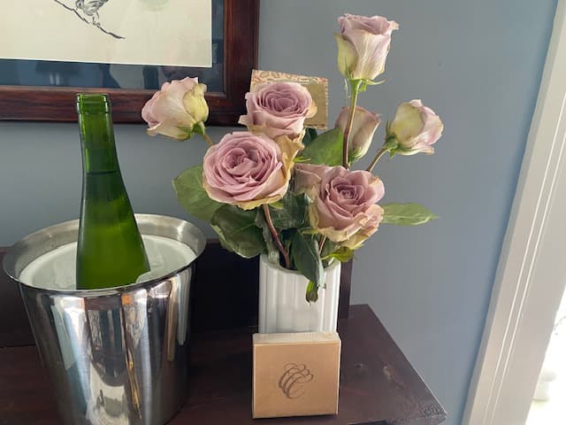 A green bottle stands in an ice bucket beside a vase of pink roses and a small gift box on a wooden table.