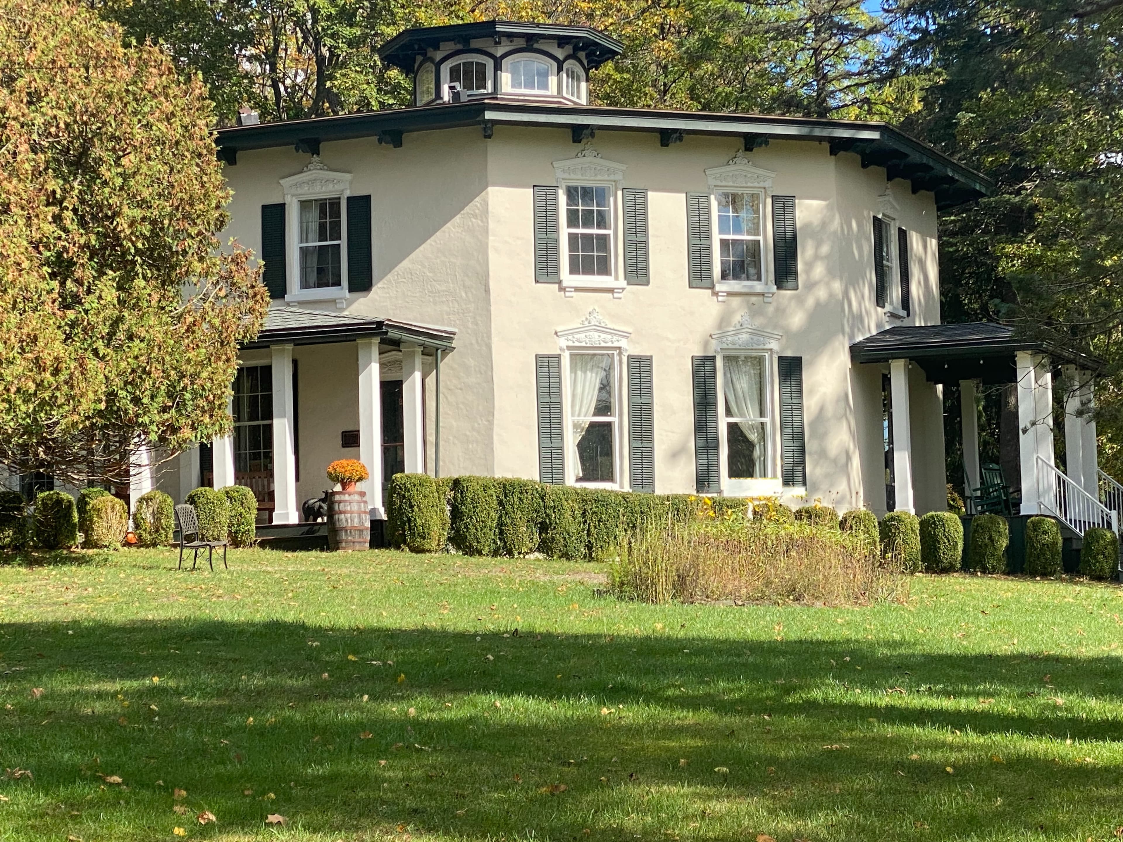 A two-story, round house with green shutters and a porch surrounded by manicured hedges and trees. A two-story, round house with green shutters and a porch surrounded by manicured hedges and trees.